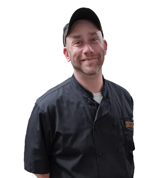 A person wearing a black chef’s coat, a dark cap, and a name tag, smiling.