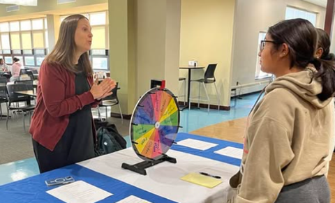 Two people stand at a table in a bright room with a colorful prize wheel placed between them.