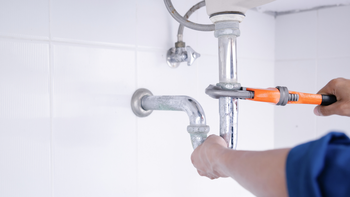 Plumber tightening under-sink pipes with orange-handled wrench in a white tiled bathroom sink area