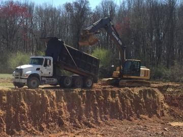 Excavator loading dirt into a white dump truck on a wooded construction site
