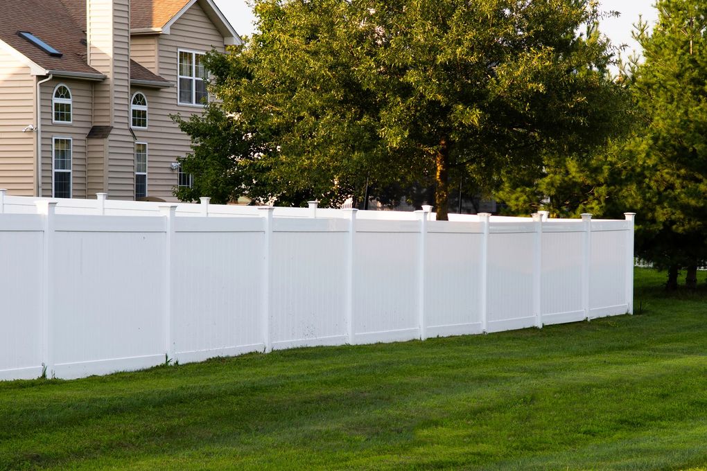 A white vinyl privacy fence stands in a green yard in front of a tan house and a large tree.