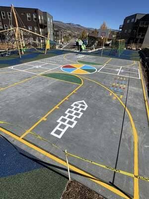 Outdoor playground court with colorful hopscotch markings and a large four-color circle, surrounded by buildings.