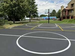 Outdoor basketball court with white circles and yellow lines near trees and buildings under a cloudy sky