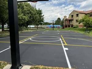 Empty outdoor basketball court with white and yellow lines beside apartment buildings and trees