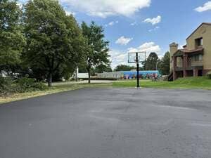 Empty paved court beside a brick building, with trees and a distant basketball hoop under a cloudy sky