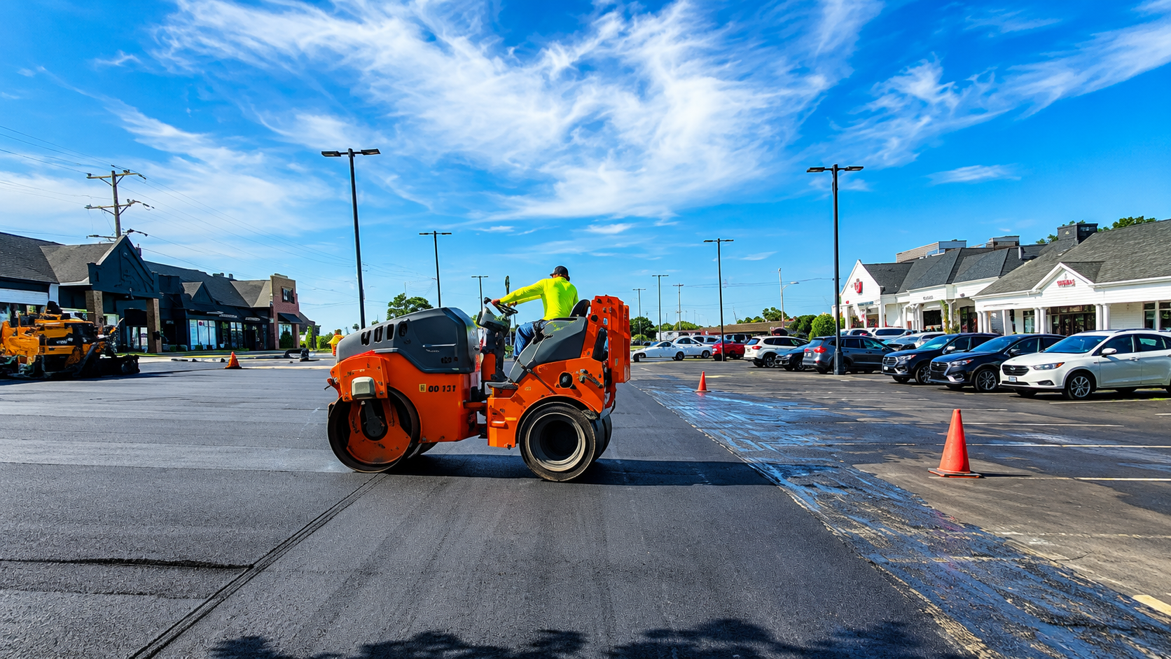 Orange road roller paving a parking lot with workers nearby and parked cars in the background