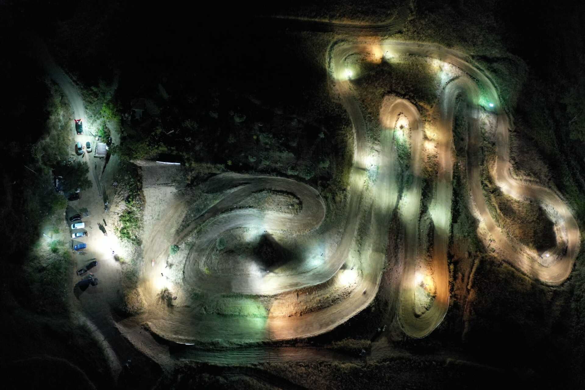 Vista aerea notturna di una pista tortuosa illuminata da fari e riflettori, con auto parcheggiate ai lati.