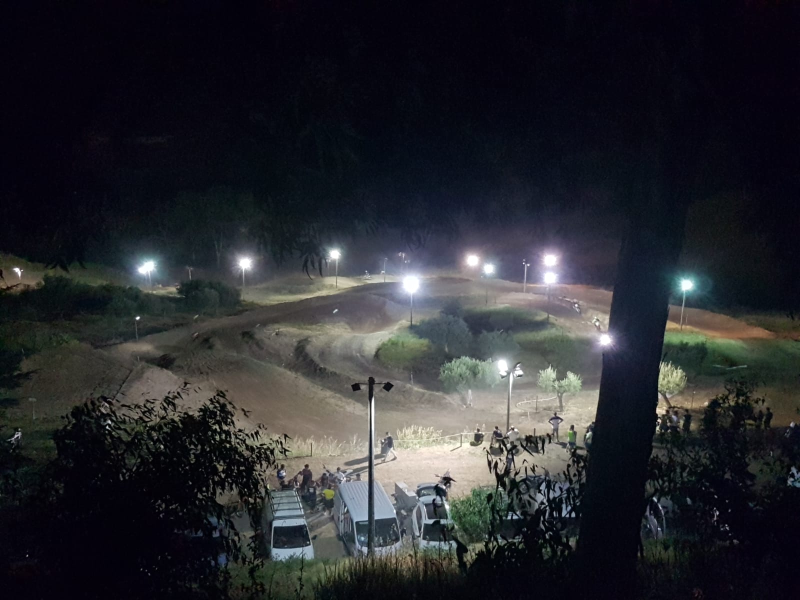 Pista notturna per moto da cross illuminata da riflettori. Spettatori e veicoli visibili.