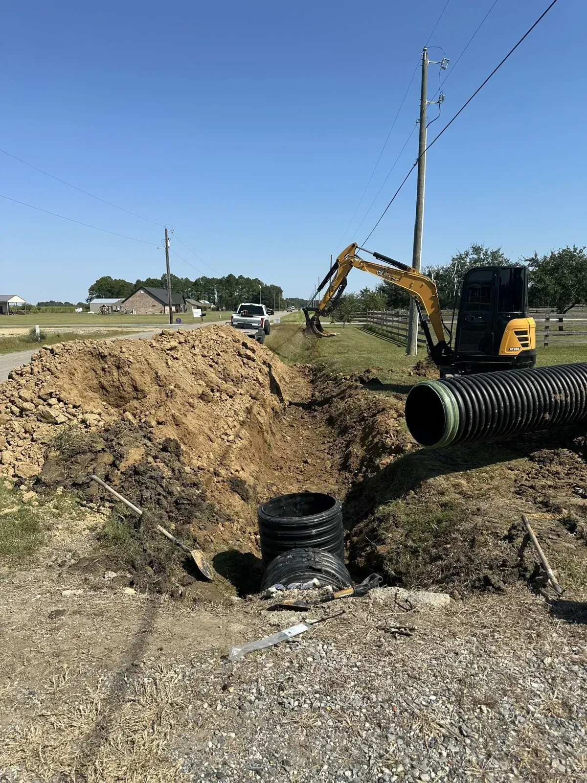 Yellow mini excavator digging a ditch trench and placing corrugated culvert drainage pipe along a roadside property in Louisiana.