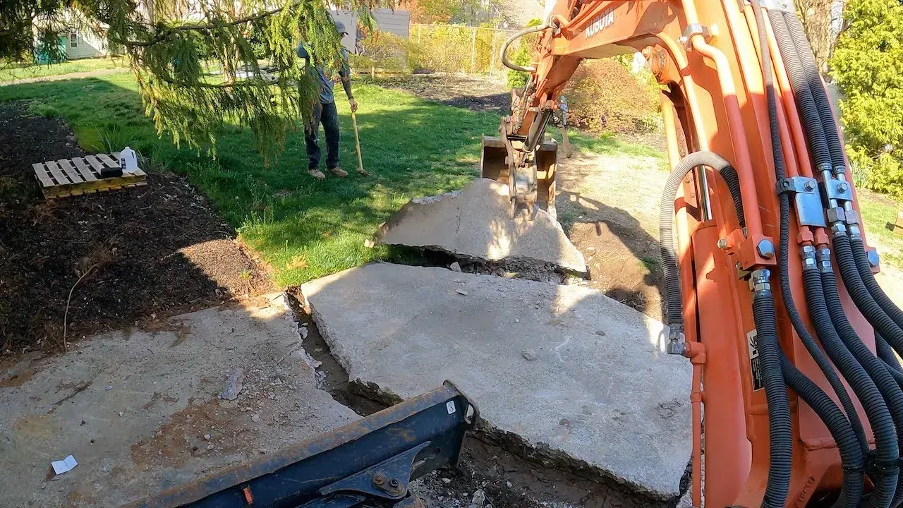 Excavator breaking up and removing a concrete slab in a residential yard