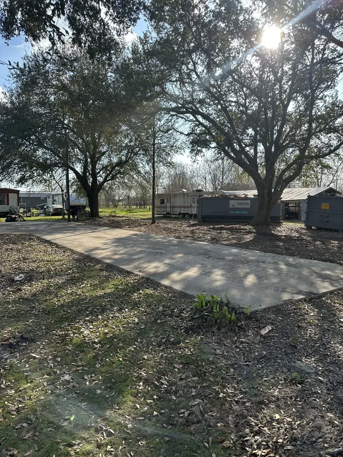 Cleared demolition site with a concrete pad beneath mature oak trees and a roll-off dumpster in the background in New Iberia, LA.
