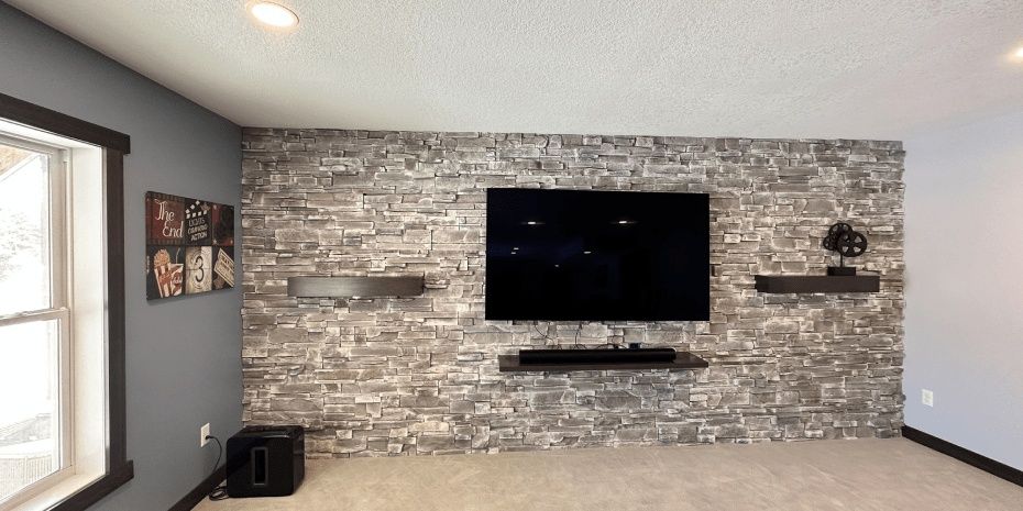 Modern living room with stone accent wall, wall-mounted TV, floating shelves, and gray carpeted floor.
