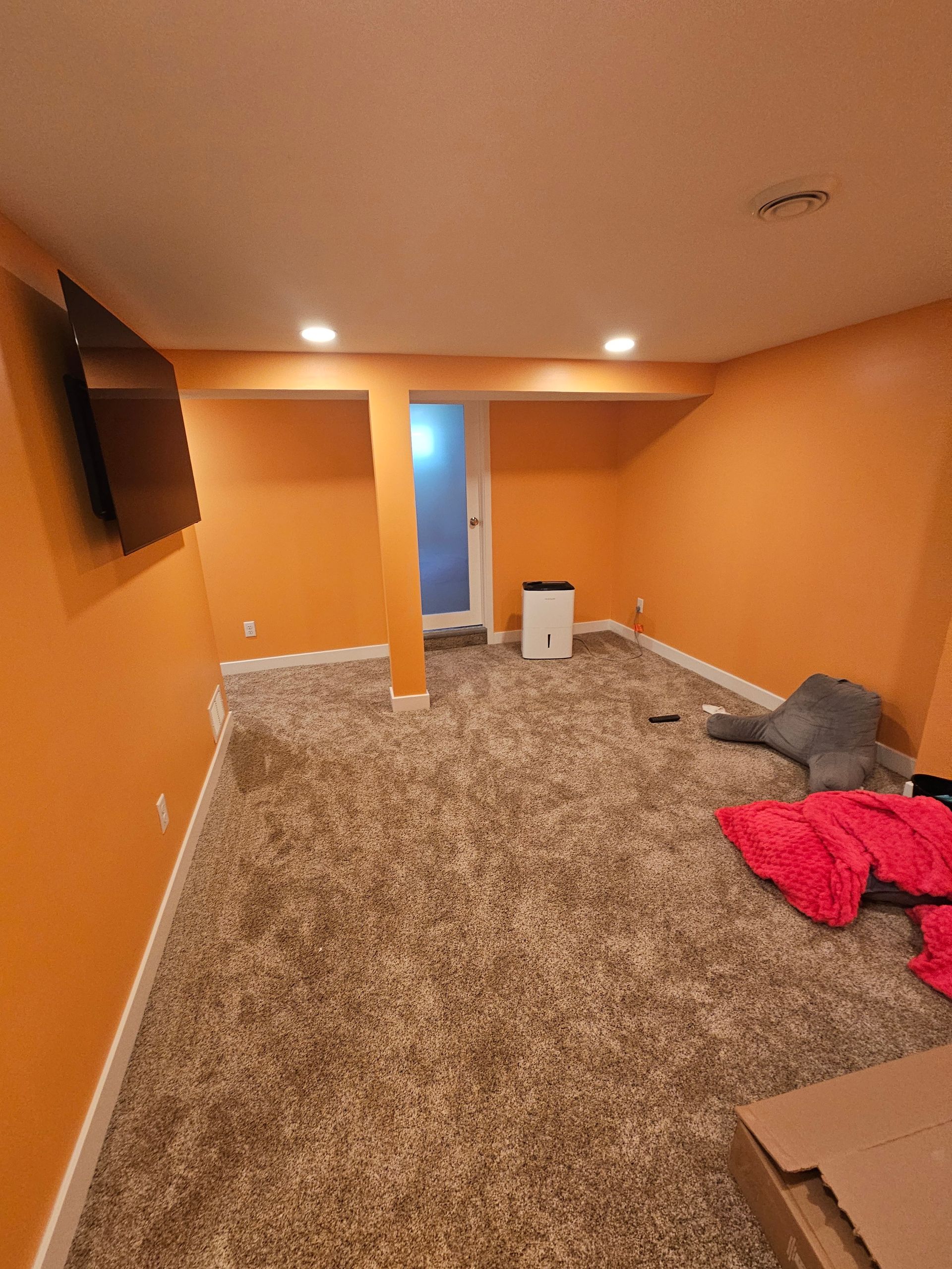 Orange basement room with wall-mounted TV, carpeted floor, and a red blanket on the floor.