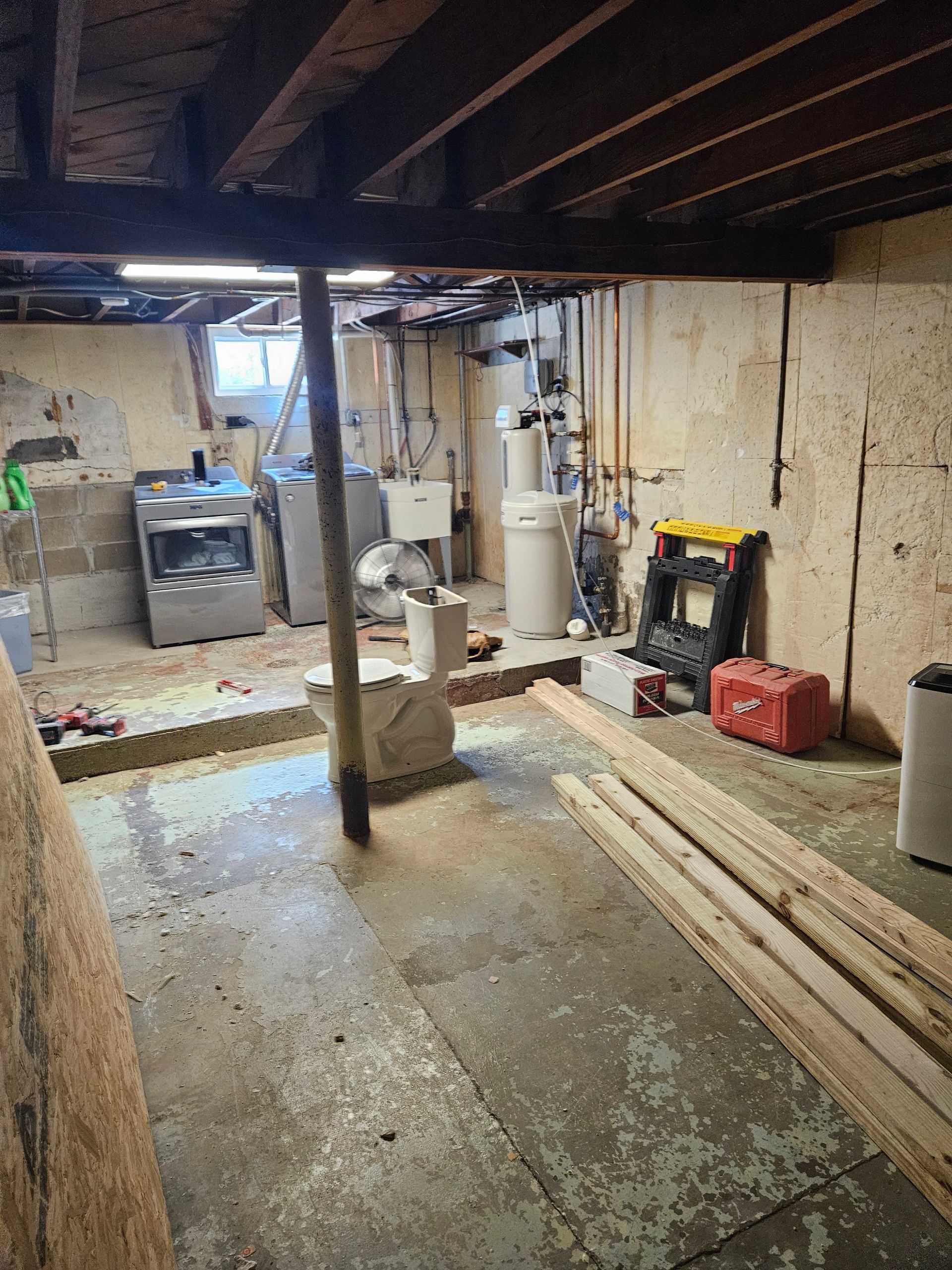 Basement under renovation with exposed beams, concrete floor, tools, and lumber stacked along the wall