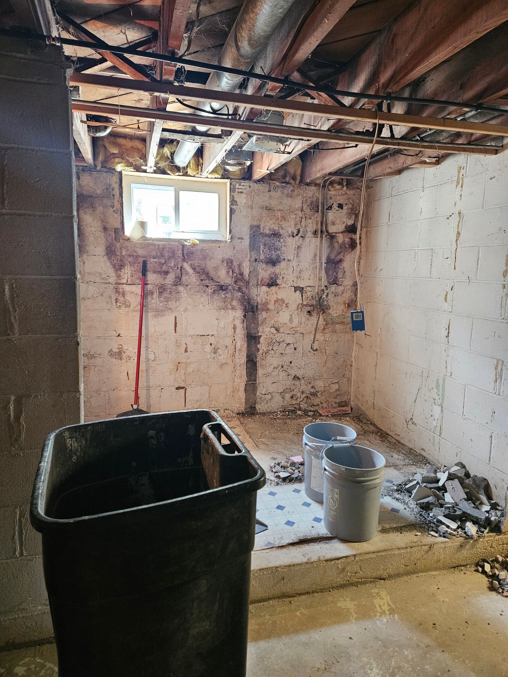 Basement room under renovation with exposed beams, small window, buckets, and debris on the floor