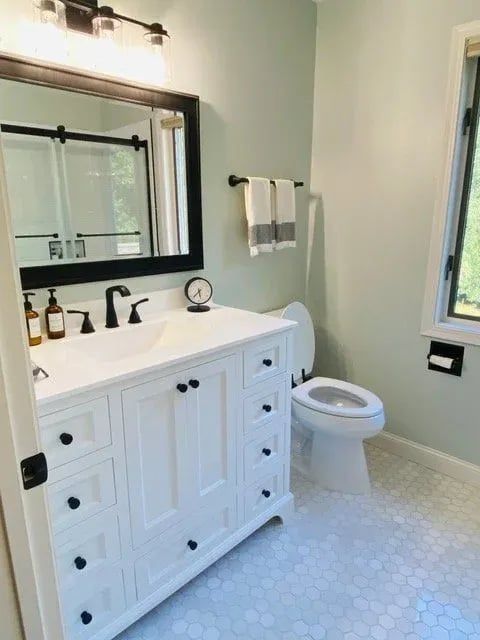 Bright bathroom with white vanity, black fixtures, wall mirror, and toilet on hex tile floor.