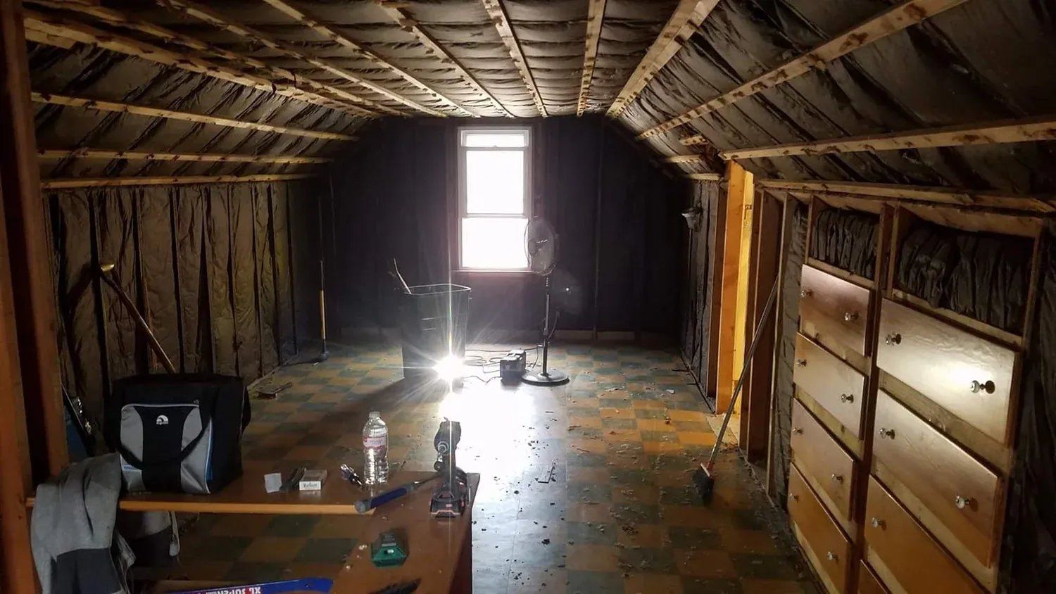 Attic interior with exposed beams, cluttered floor, and bright light from a small window