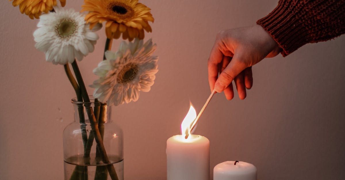 Een hand steekt een witte kaars aan naast bloemen in een vaas, een warme binnenomgeving.