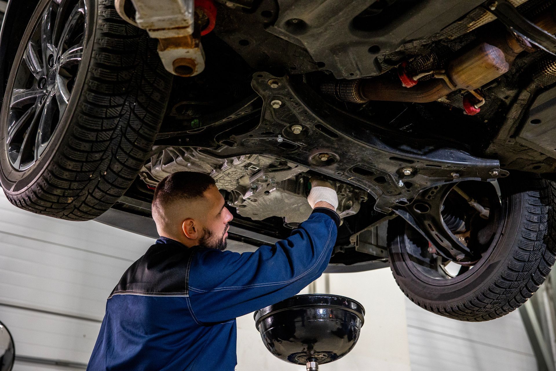 A mechanic performing an oil change under a raised car with a drain pan.