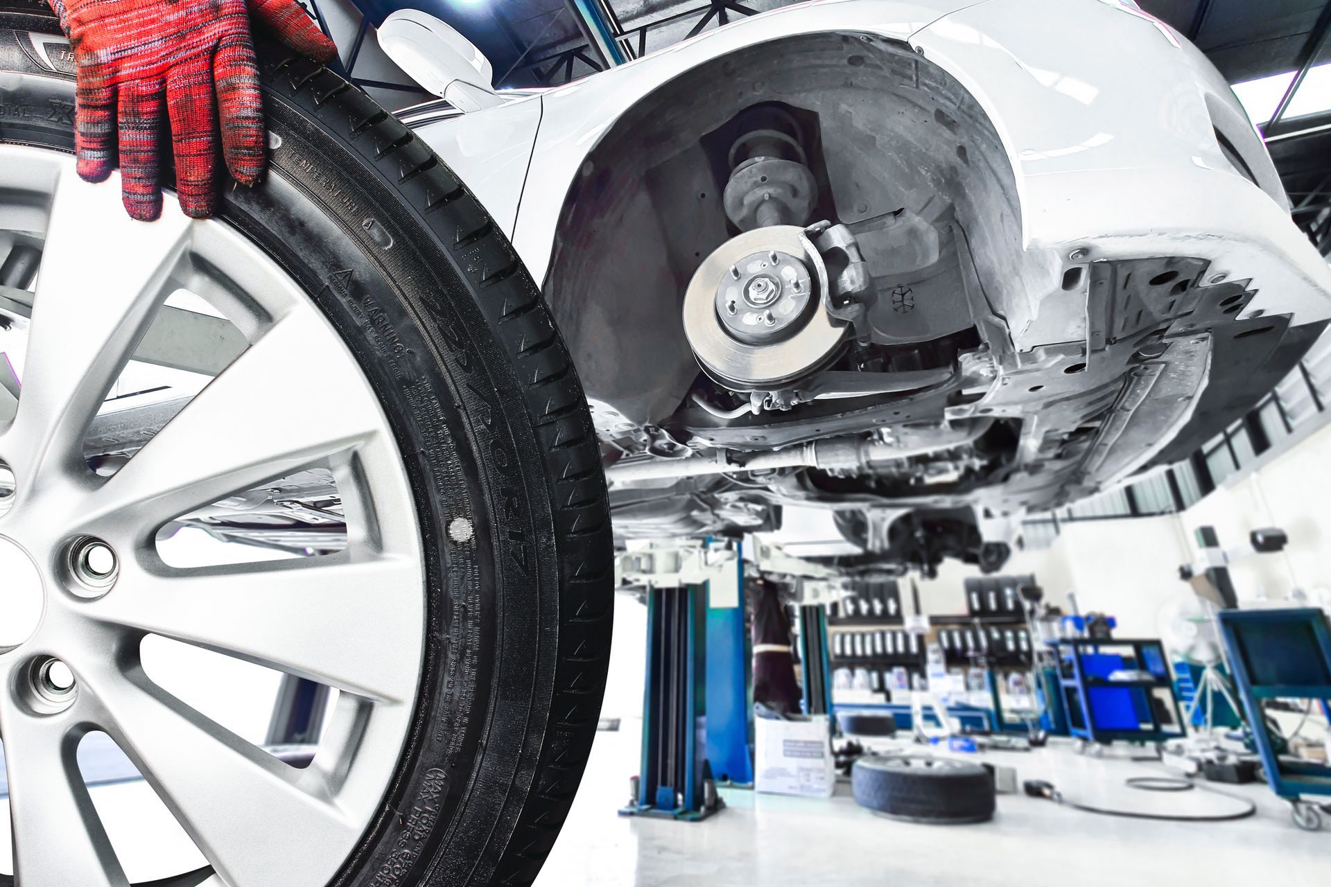 A mechanic's hand holds a car wheel removed from a white car elevated on a lift. A mechanic's hand holds a car wheel removed from a white car elevated on a lift.