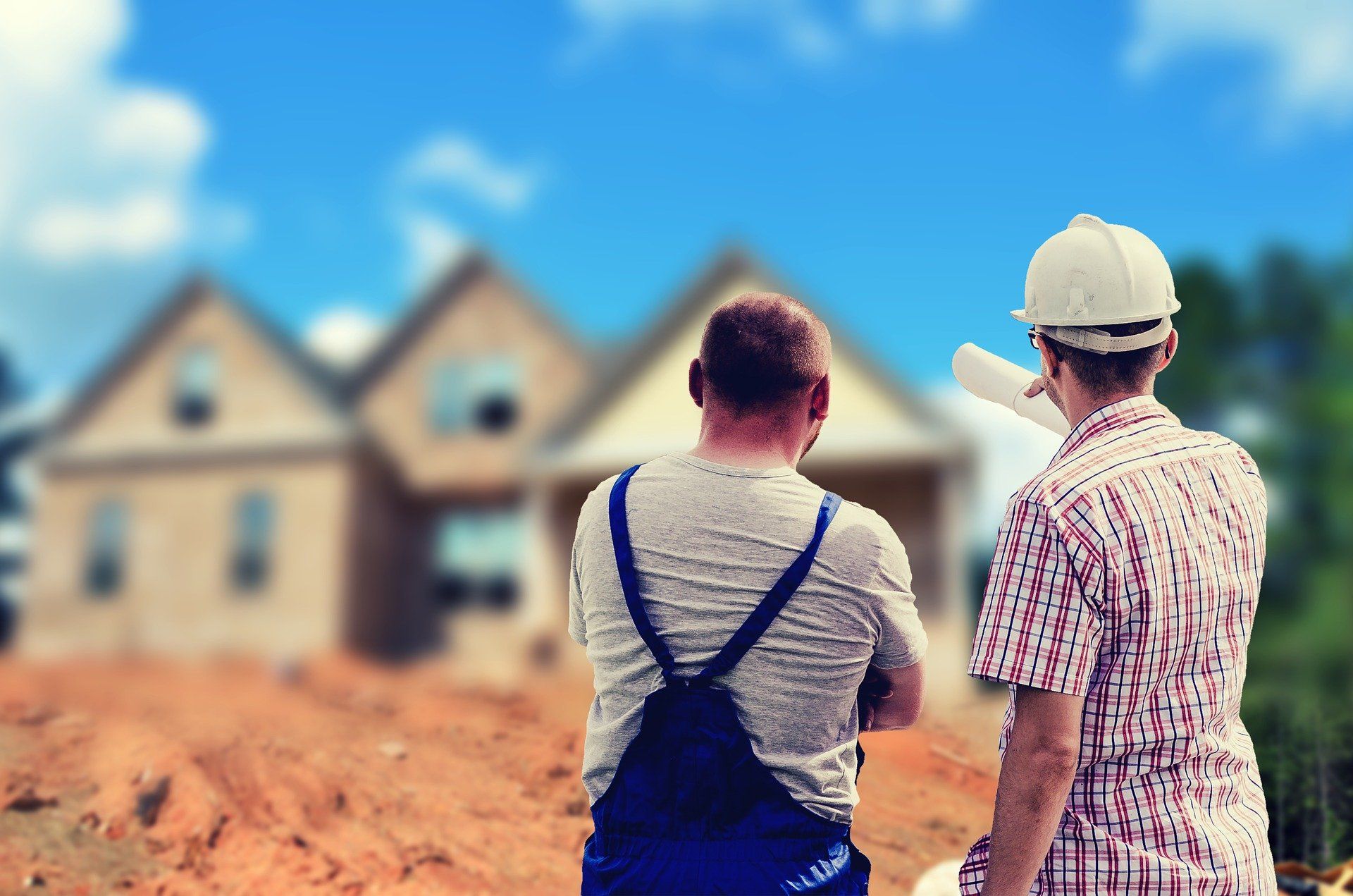 Two construction workers looking at a house under construction; one holds plans.