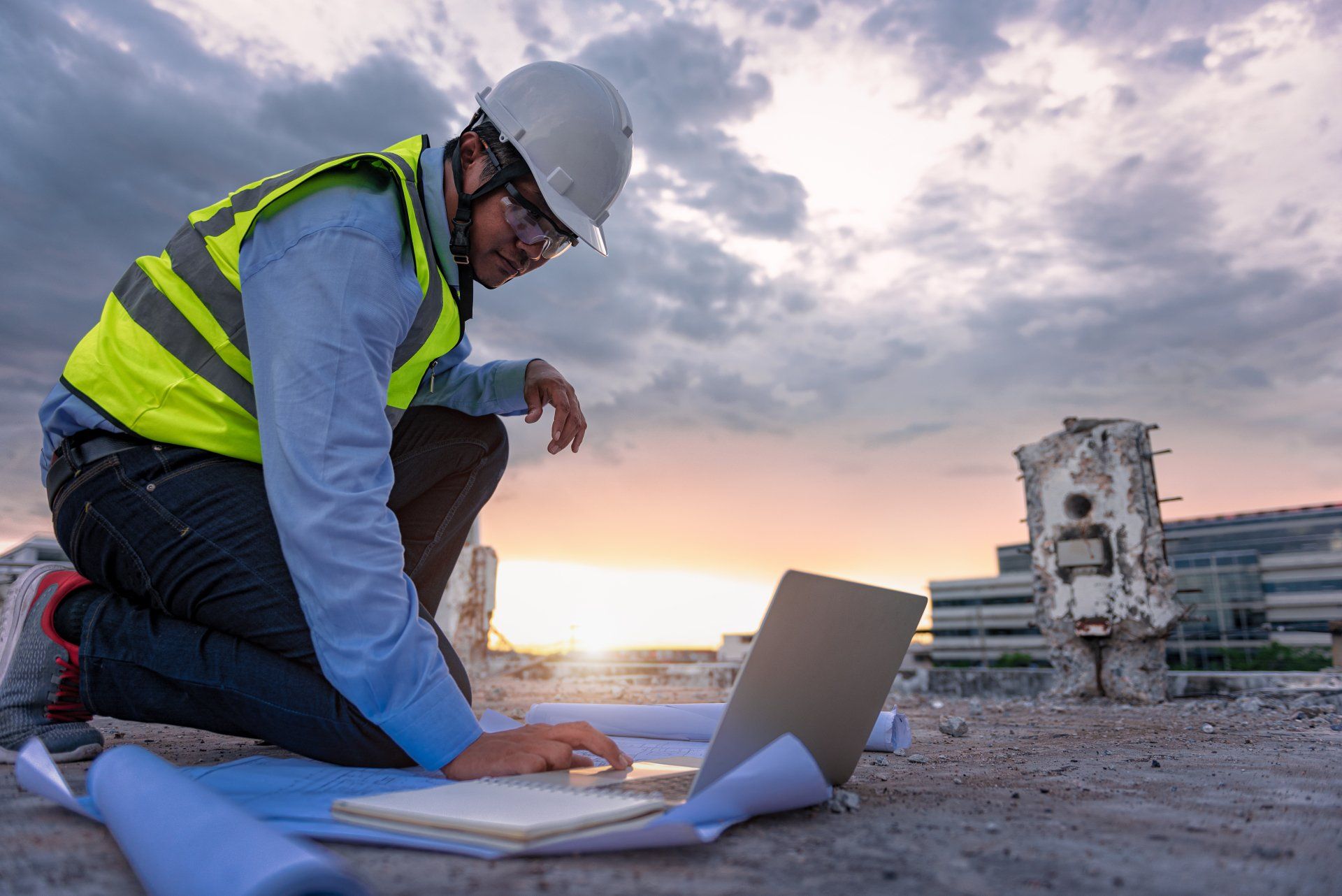 Un ouvrier du bâtiment, agenouillé sur un toit au coucher du soleil, regarde un ordinateur portable et des plans. Il porte un casque et un gilet de sécurité.