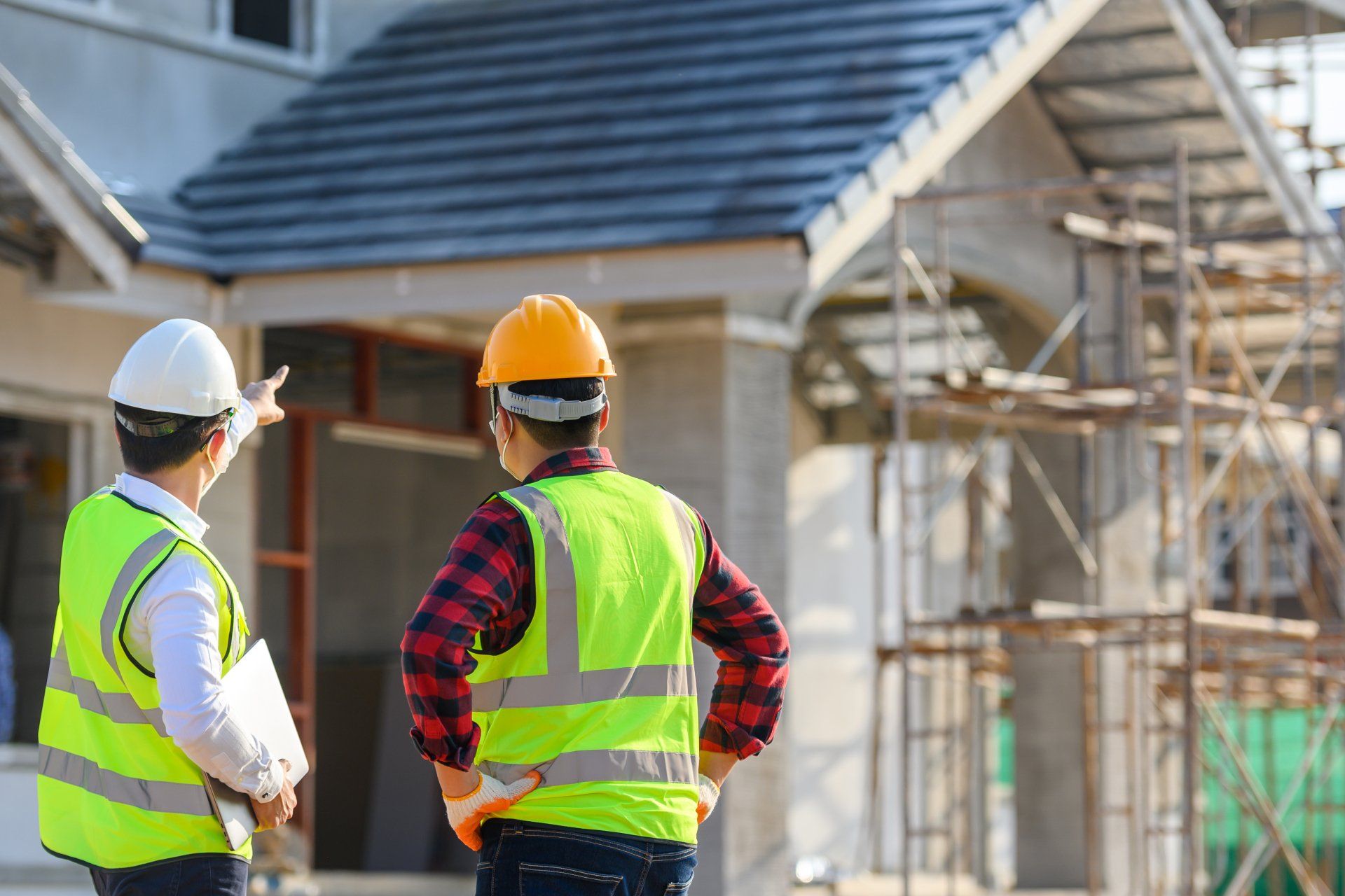 Two construction workers in safety vests and hard hats discuss a building project outdoors.