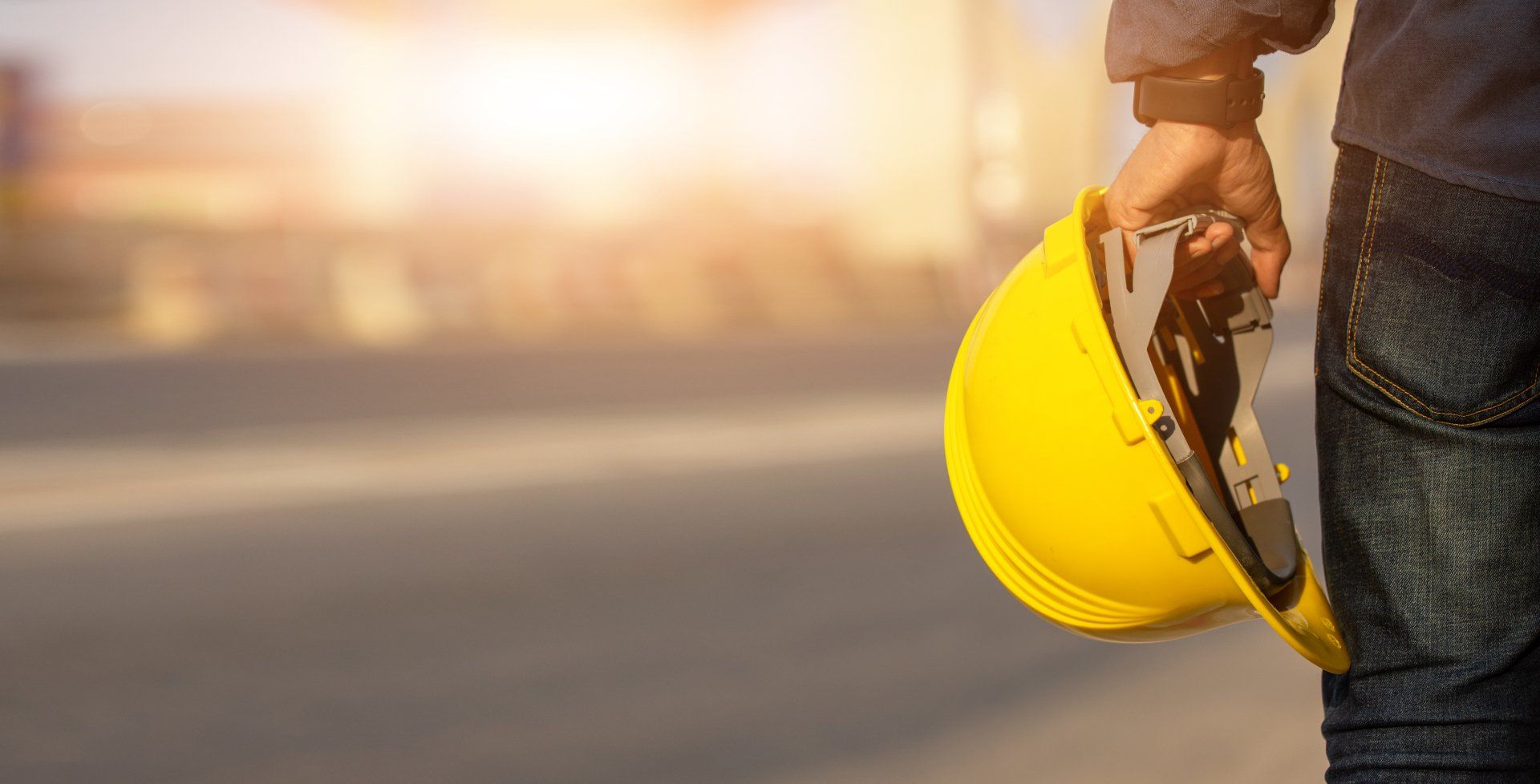 Personne tenant un casque de chantier jaune à l'extérieur, lumière du soleil en arrière-plan.