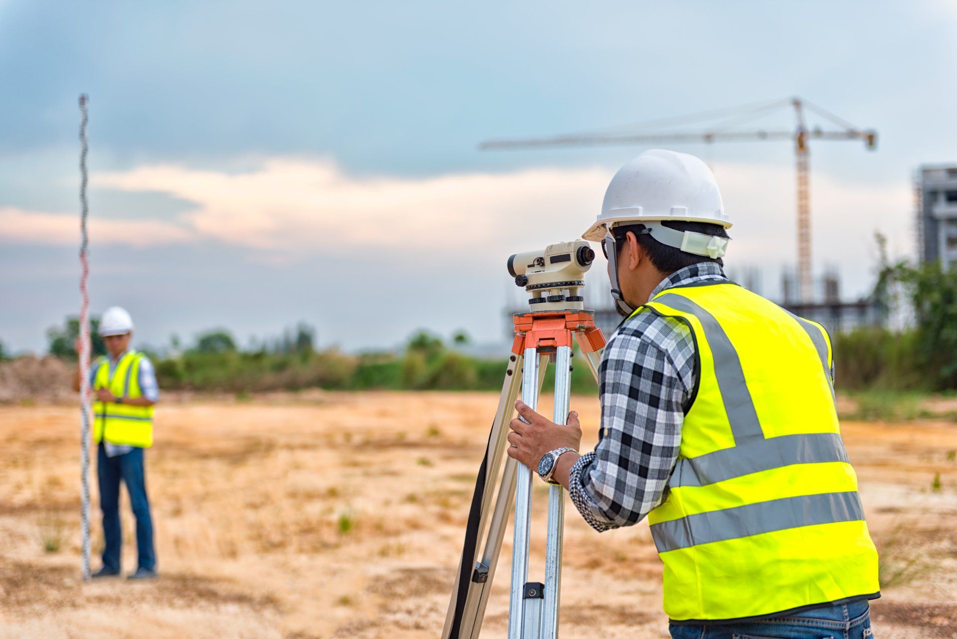 Two surveyors on a construction site using surveying equipment. One looks through a level, the other holds a surveying rod.