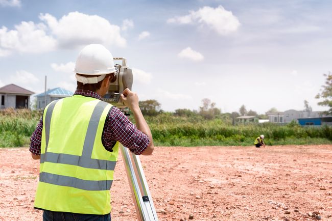 Géomètre utilisant du matériel sur un chantier de construction ; portant un casque et un gilet de sécurité.