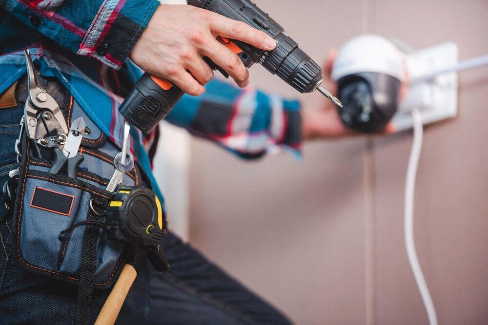 A Man is Installing a Security Camera With a Drill — South Coast Locksmiths in Fairy Meadow, NSW