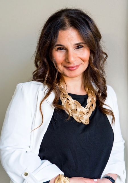 Woman in white blazer and gold necklace smiling, in front of neutral wall.