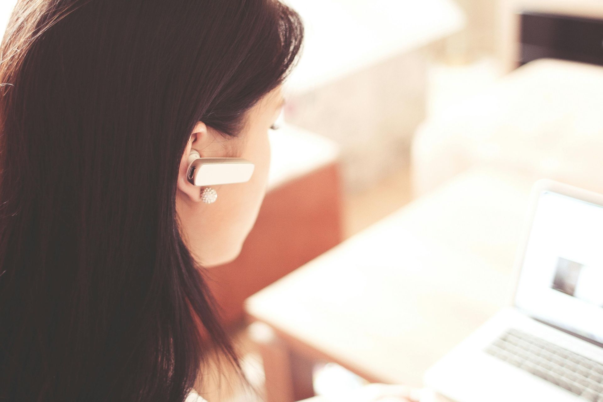 Woman with Bluetooth headset looking at a laptop screen at a desk.