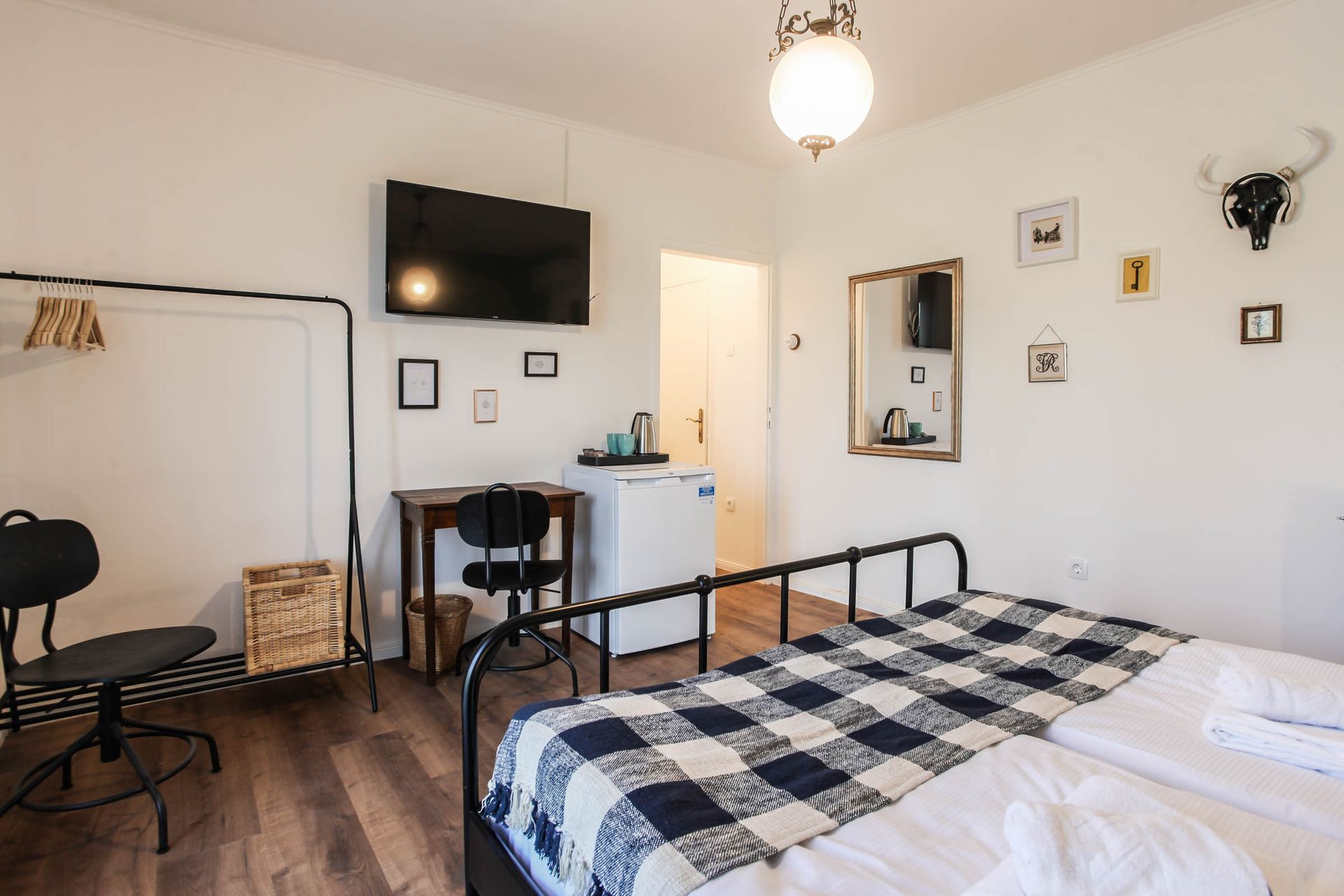 Cozy bedroom with black metal bed, checkered blanket, wall TV, desk, and white walls.