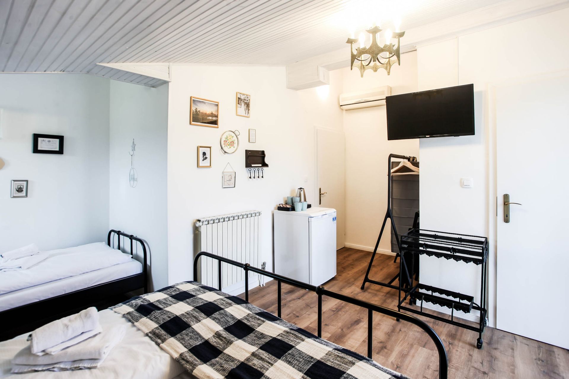Small bedroom with two beds, checkered blankets, white walls, TV, and a chandelier.