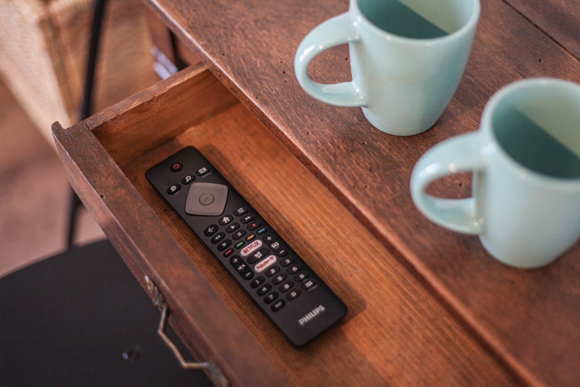 Black remote control in a wooden drawer beside two light blue mugs on a tray