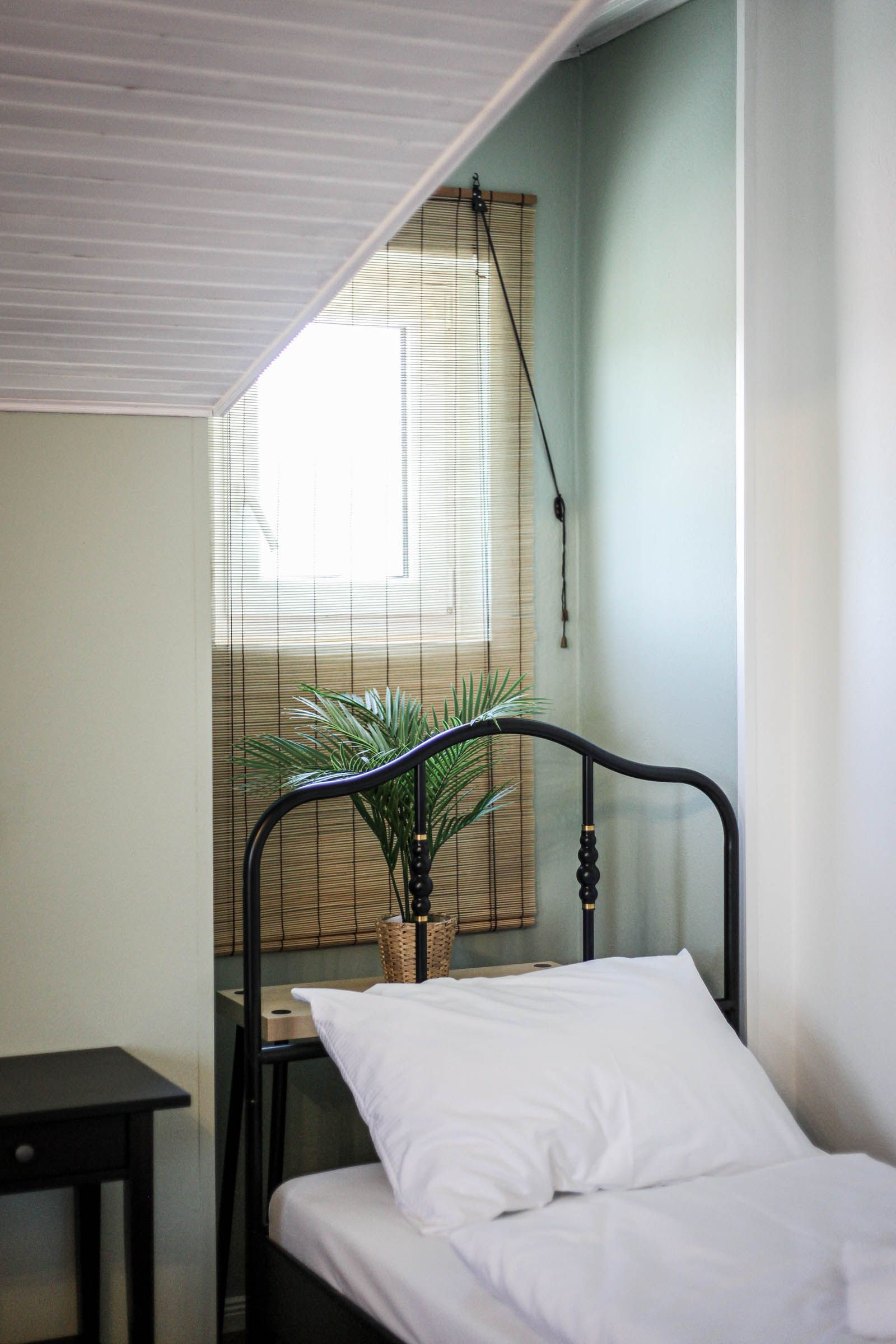 Small bedroom nook with a metal bed, white bedding, and a window with blinds and bamboo shades.