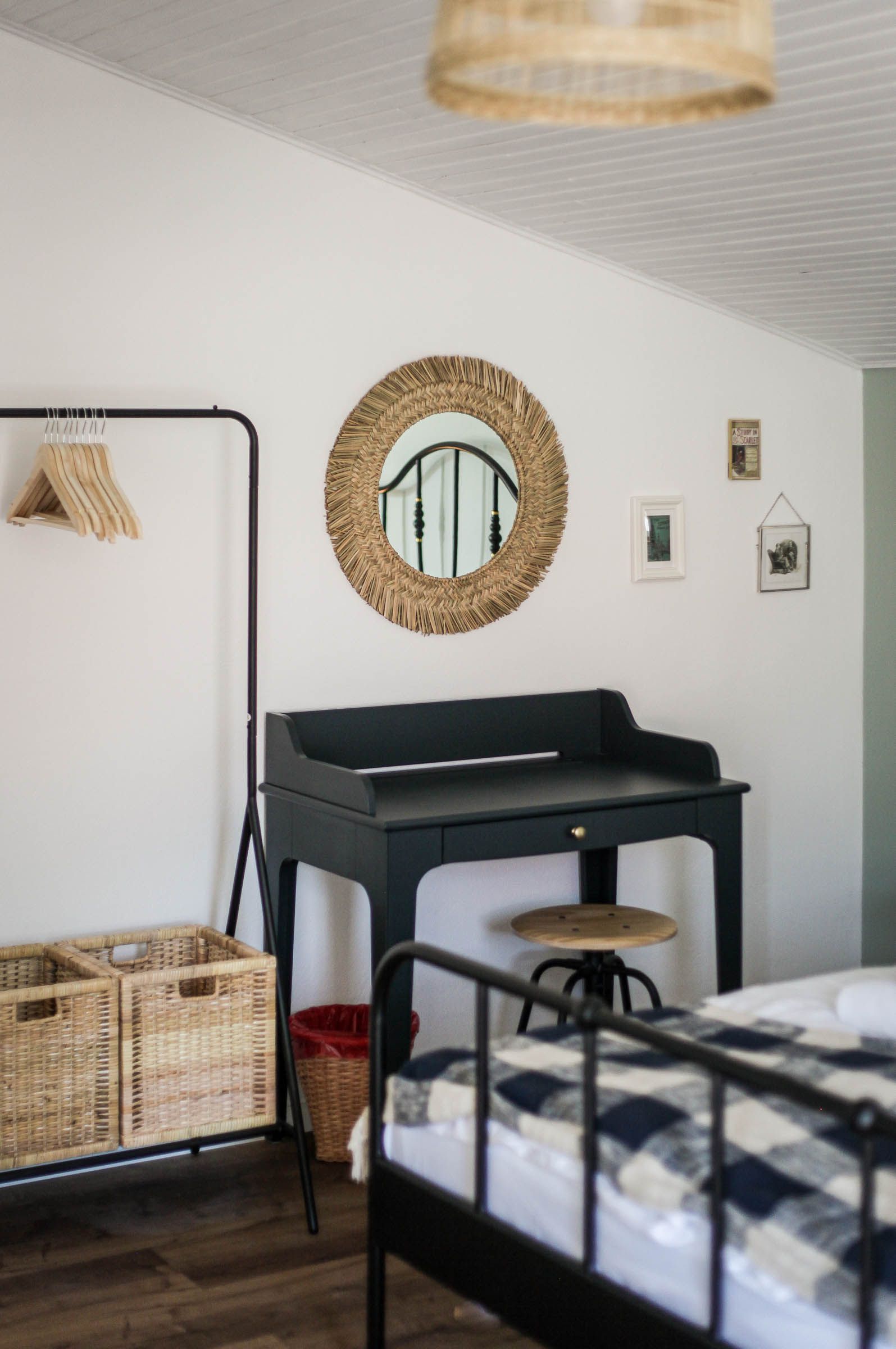 Cozy bedroom with black desk, woven mirror, framed wall art, wicker baskets, and plaid bedding.