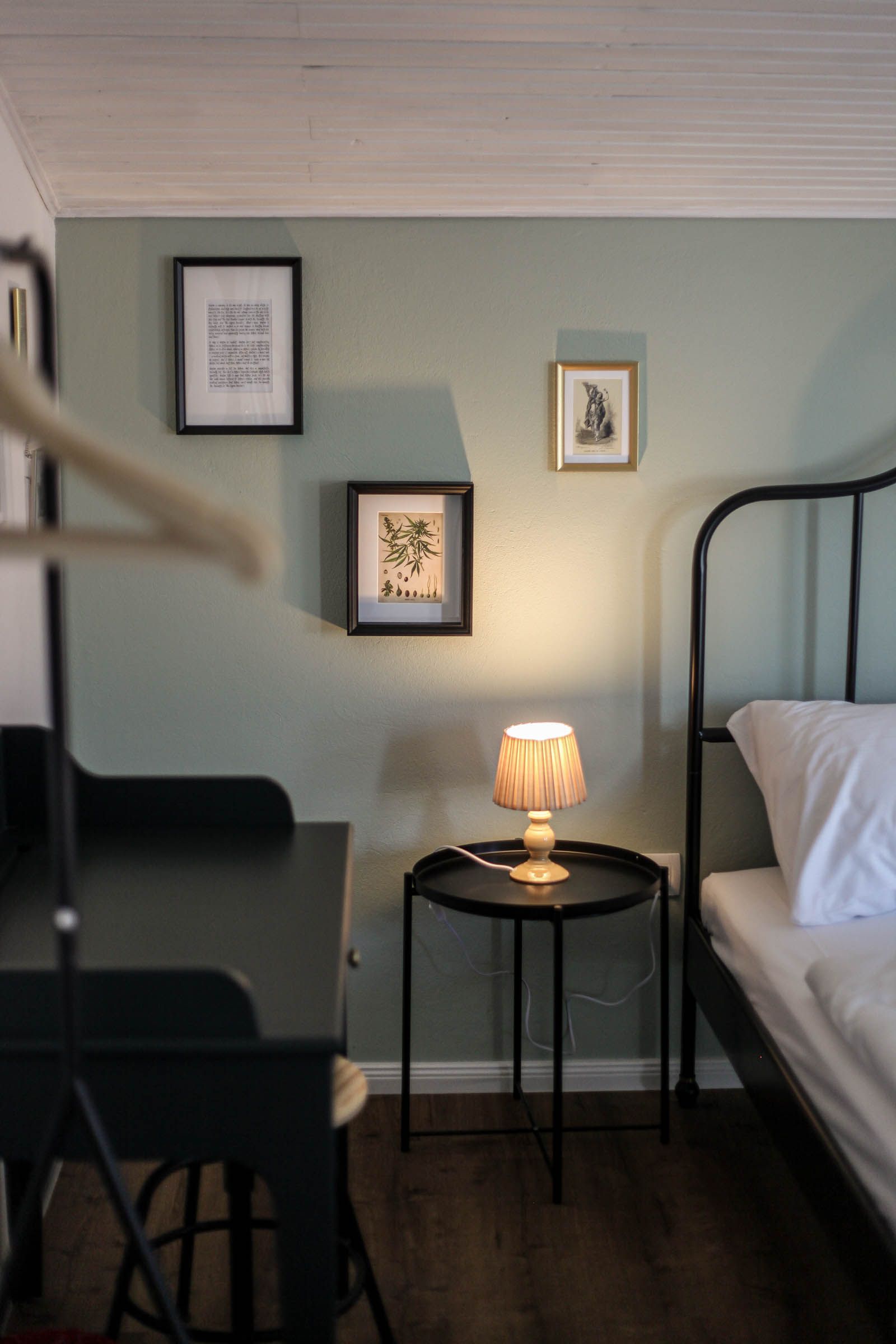 Cozy bedroom corner with black metal bed, lit bedside lamp, framed art, and a small round table.