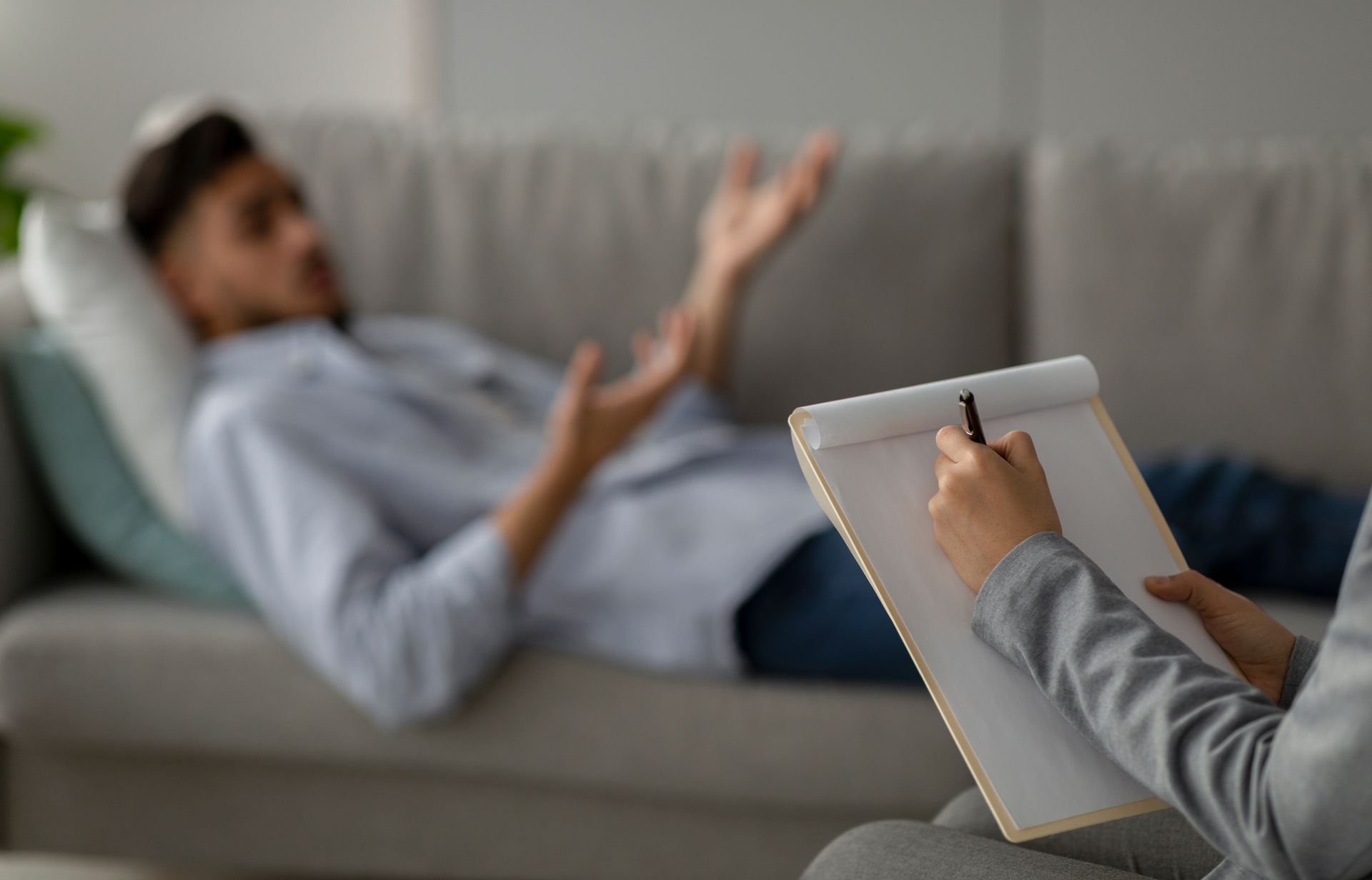 Client reclining on a couch gesturing while explaining feelings, with a therapist's hand taking note