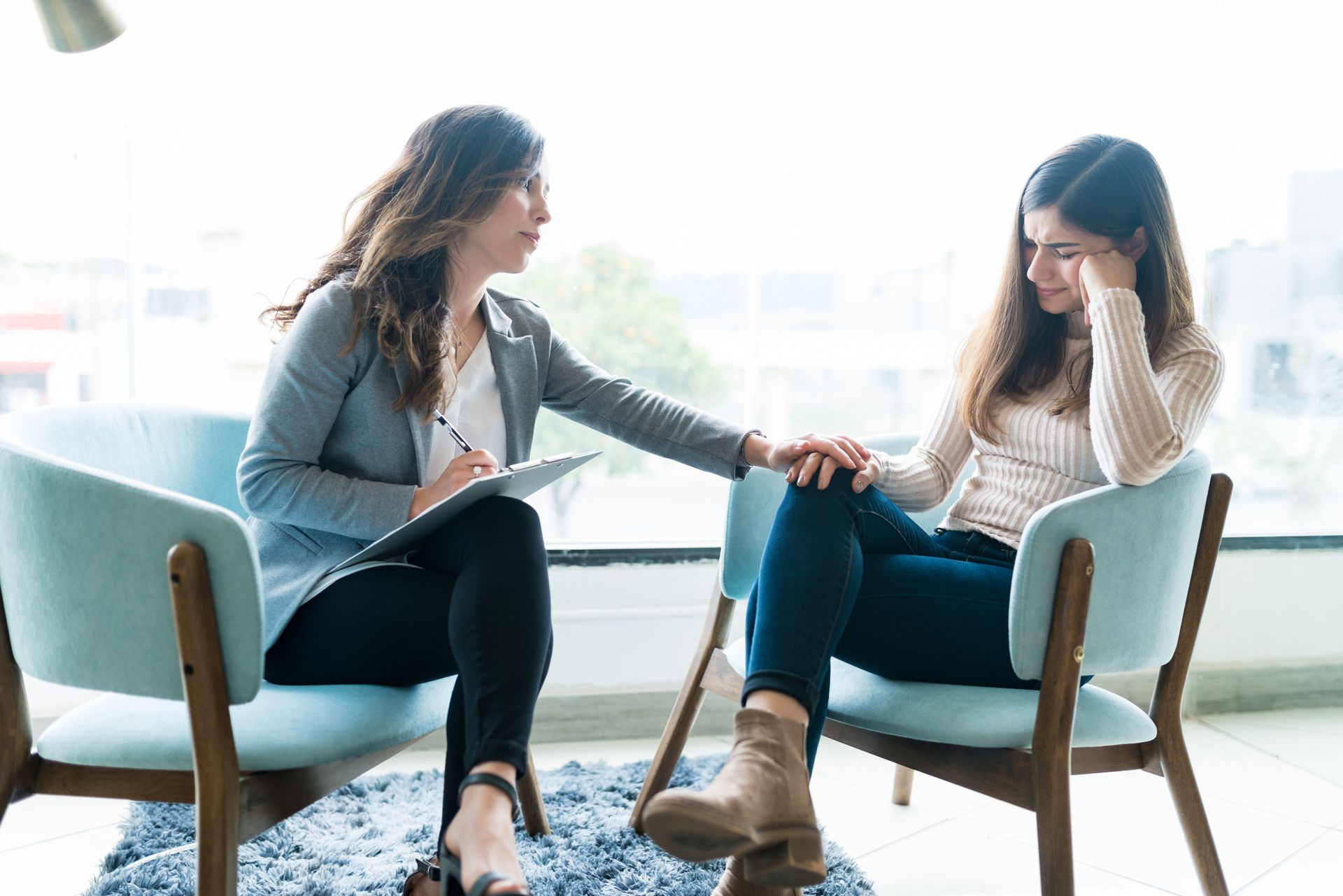 Therapist taking notes while speaking with a seated client in a bright office Two women sit in armchairs, one comforting the other by holding her hand in a bright room.