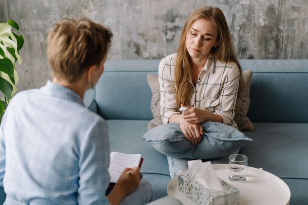 Therapist talking with a client in a bright counseling room, seated on blue and white furniture.