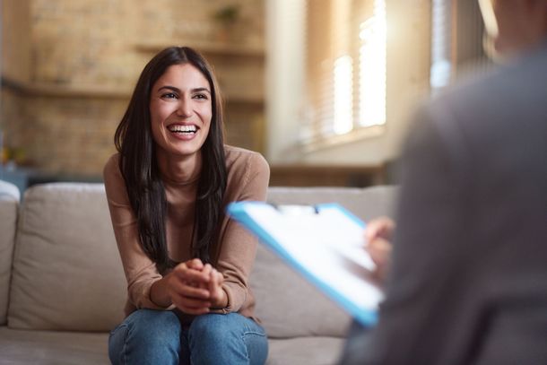 Smiling woman in a living room, speaking with a person holding a clipboard.