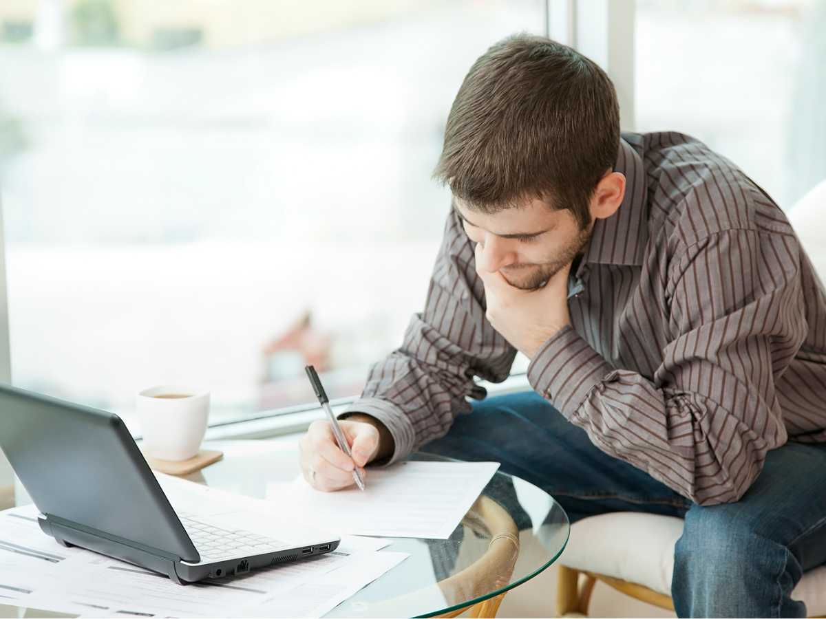 A man is sitting at a table completing forms with a laptop open in front of him