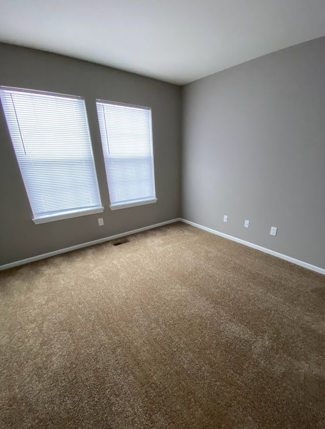 An empty bedroom with a carpeted floor and two windows.