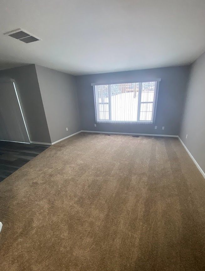 An empty living room with a large window and brown carpet.