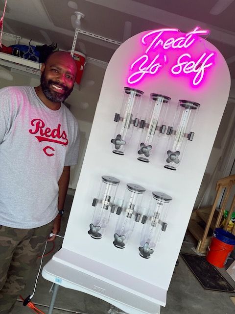 Man standing next to a white dispenser with six clear canisters; pink neon sign says 
