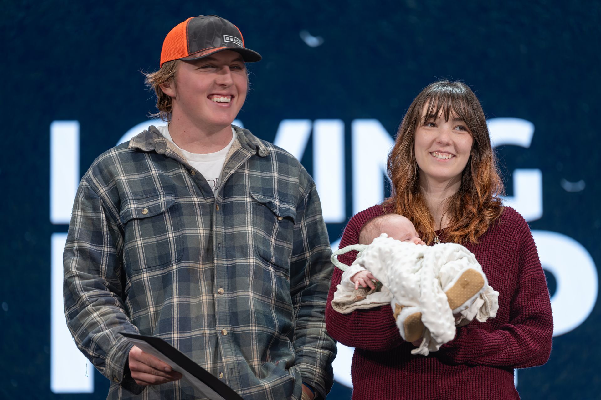 Two people onstage smiling, one holding a white goat in a maroon sweater, with a large blurred sign behind them