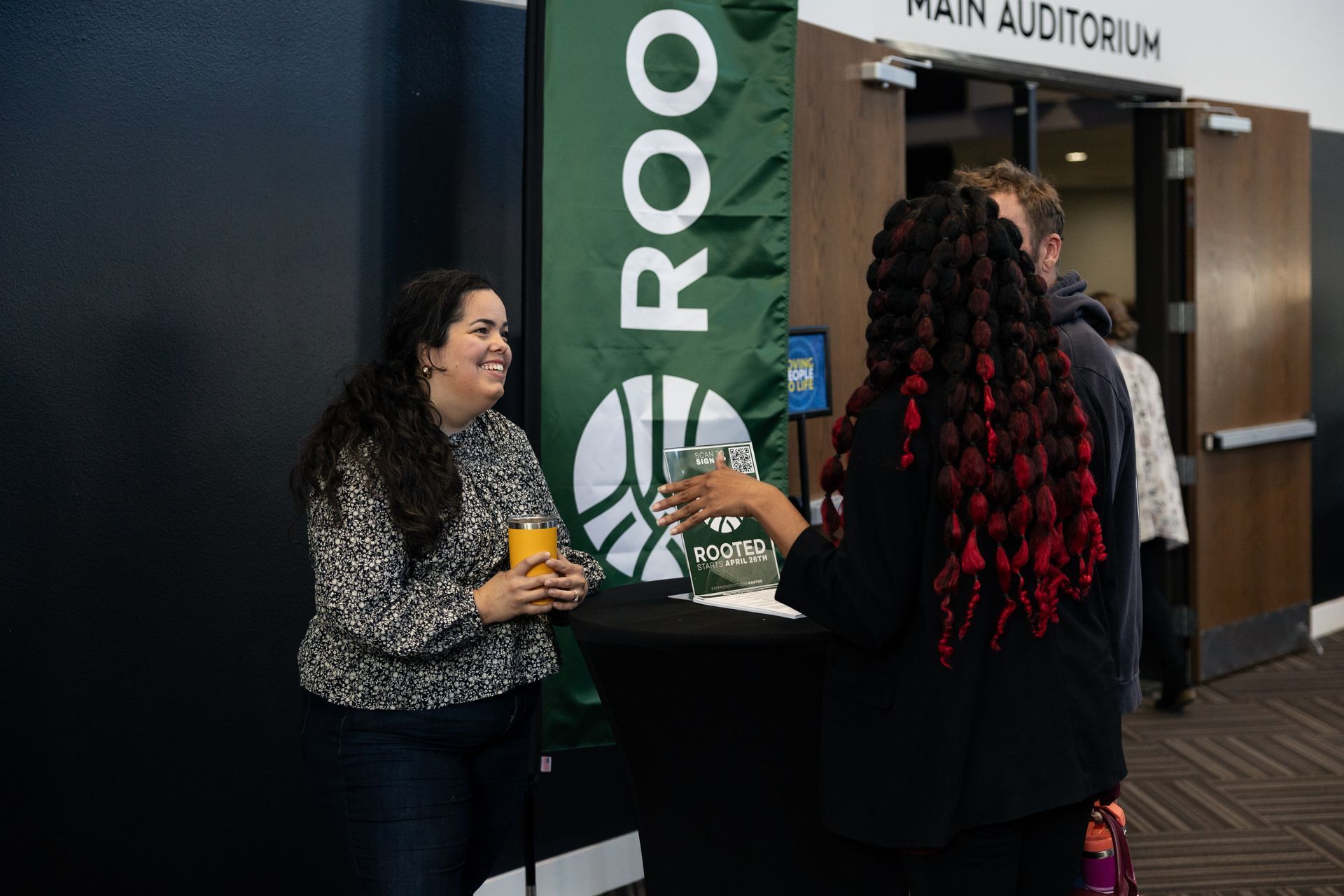Two people talking at a reception table near a “ROOM” sign and the Main Auditorium entrance.
