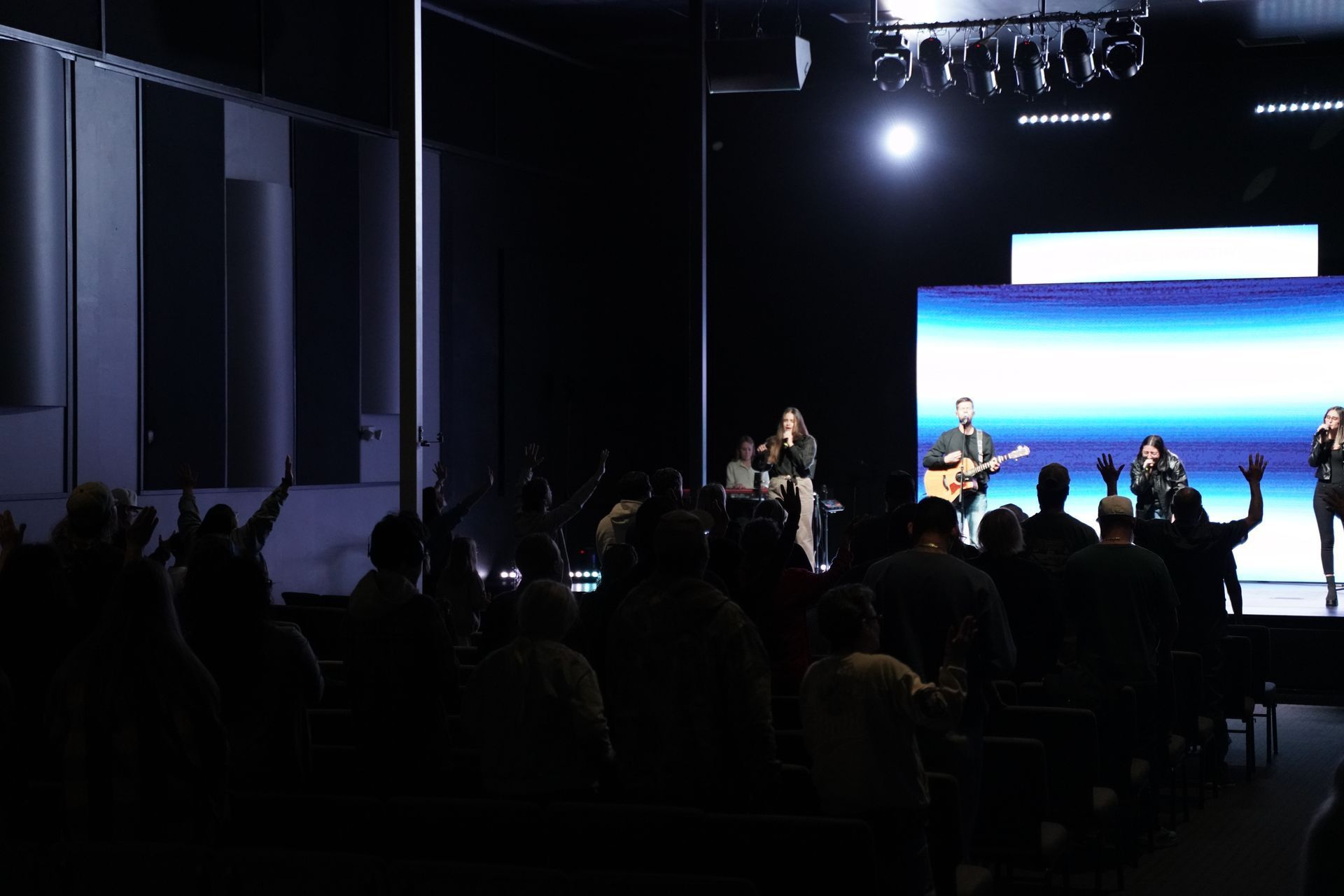 Live band performing on a dark stage with a bright blue ocean backdrop and audience silhouettes